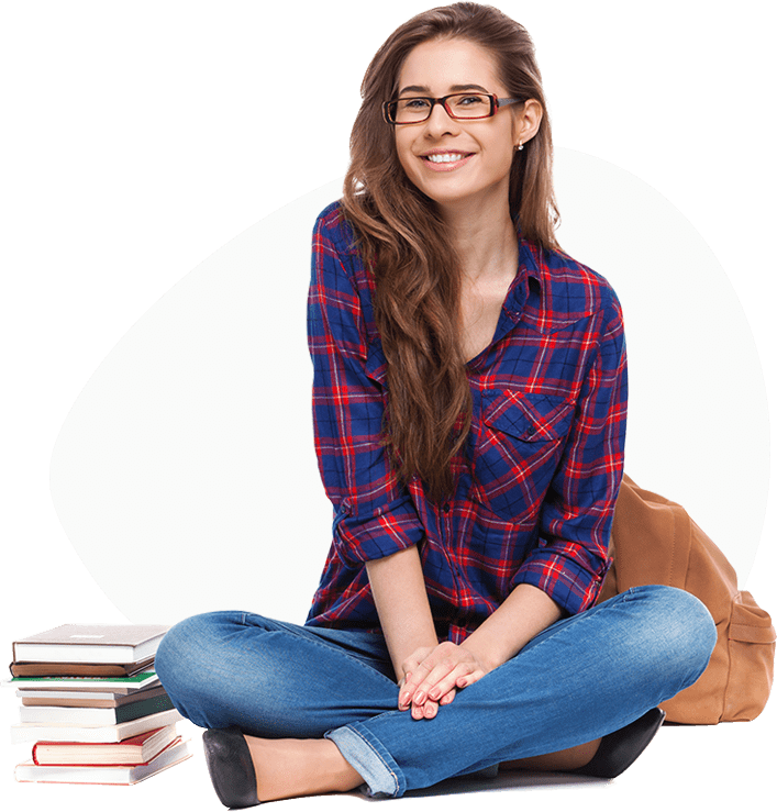 A woman sitting crossed legged on the floor. She is wearing blue jeans with black pumps. Her hands are folded over each other on her calve. She is wearing a black long sleeved top with small red squares which are four at a time to form blocks. She has long dark, wavy hair and wears glasses. She is also wearing earrings. She is smiling. Next to her, is a pile of books. .