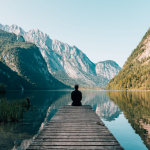 Person sitting at the end of a pier in a lake with mountains on either side, the one side in sunlight, the other side in shade, representing goal setting with Ignite Co