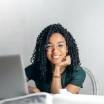 Young African lady sitting at a desk behind a laptop with her head resting on her hand while she;s smiling