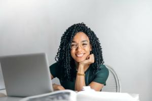 Young African lady sitting at a desk behind a laptop with her head resting on her hand while she;s smiling
