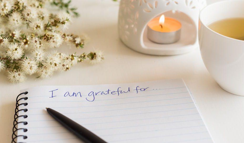 Notebook with the words written "I am grateful" and a pen on it, with white flowers on the left, and a candle and cup with tea on the right of it